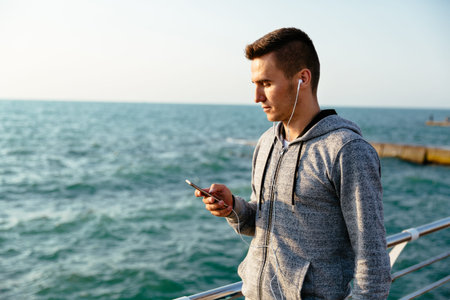 Handsome pensive man in sport jacket and headphones, looking at mobile phone screen, during walking...