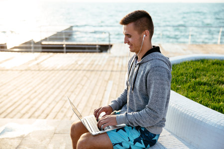 Happy young attractive man in headphones, working on laptop, sitting on bench, on quay, near the sea.の写真素材