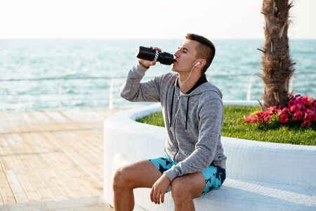 Profile of handsome sportsman in sportswear drinking a water from bottle after workout, sitting on bench, on quay, near the ocean.の写真素材