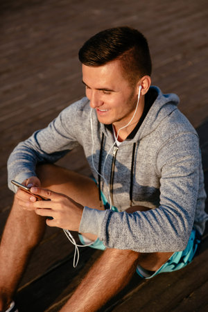 Joyful handsome sportsman in earphones, listening to music, while using a mobile phone, resting...