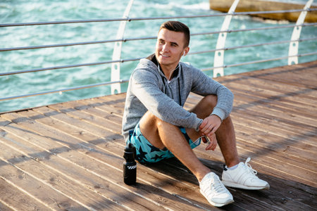 Young stylish sportsman listening to music in earphones, resting on pier after workout, with bottle...