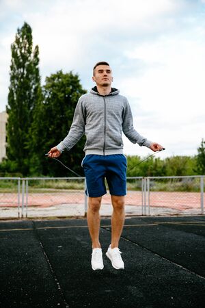 Strong active guy jumps on the skipping rope in grey jacket, blue shorts, white sport shoes, outdoorsの写真素材