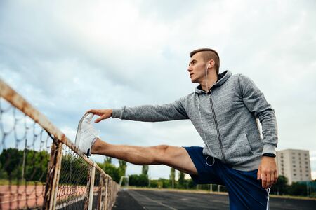 Serious sportsman guy doing exercise, raised up his leg perpendicular to the ground, listening to music, at the stadiumの写真素材