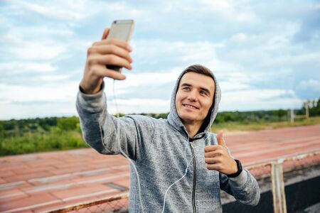 Close-up portrait of young smiling man taking selfie with smartphone, showing a thumb, wearing sweatshirt with hood, outdoorsの写真素材