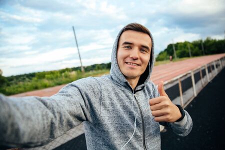 Cheerful sportive guy taking selfie, showing a thumb, listening to music, dressed in sweatshirt with hood, at the stadiumの写真素材