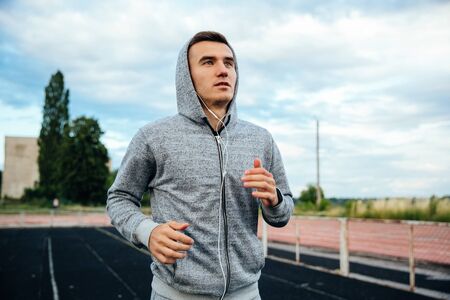 Close-up portrait of a handsome running sportsman, wearing sweatshirt with hood and listening to music, at the stadiumの写真素材