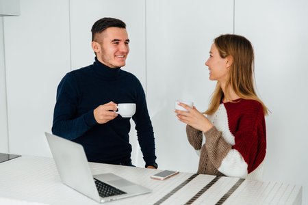 Two young smiling friends talking to each other, while drinking tea, sitting for the table with...