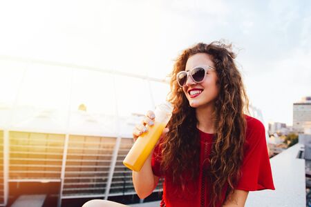 Happy young woman in sunglasses holds a bottle with fresh juice, looking away, enjoying a sunny day, outdoors.の写真素材