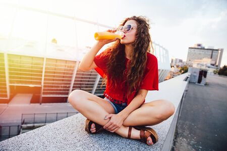 Young hipster woman with curly hair in sunglasses drinking fresh orange juice from bottle, spending time with pleasure outdoors.の写真素材