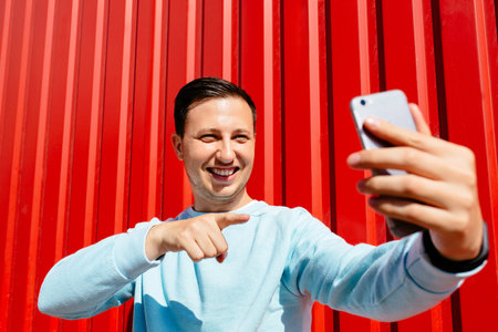 Selfie time. Young smiling man makes self portrait on smart phone on the background of red wall