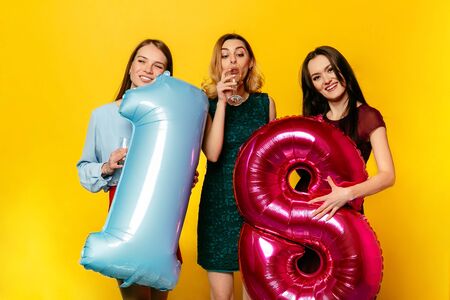 Happy birthday. Three happy pretty women in trendy dressed celebrating an anniversary with champagne, holding balloons. Isolated over yellow background.の写真素材