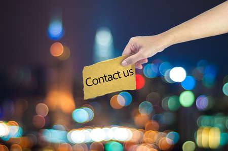 Hand holding a Contact us sign made on sugar paper with city light bokeh as backgroundの写真素材