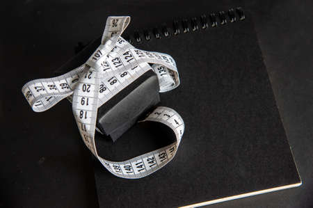 A centimeter tied with a bow on the lid of a handmade black paper gift box, which stands on a craft paper notebook. Close up. Isolated. Low key photography. Smart gift concept. Top view. Copy spaceの写真素材