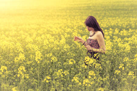 Beautiful woman in meadow of yellow flowers looking down. Attractive genuine young girl enjoying the warm summer sun in a wide green and yellow meadow. Part of series.の写真素材