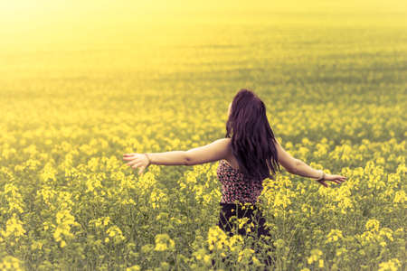 Beautiful woman in meadow of yellow flowers from behind. Attractive genuine young girl enjoying the warm summer sun in a wide green and yellow meadow. Part of series.の写真素材