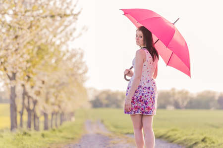 Beautiful young woman with red umbrella in spring or summer. The weather is changeable between rain and sun. The attractive girl is alone for a walk in a wonderful avenue in natureの写真素材