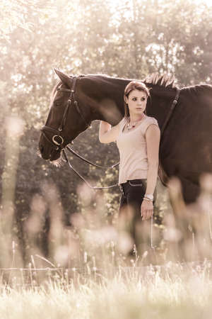 Vintage young woman equestrian with horse in summer sun Nature. Scene of a teenager horsewoman holding her best friend animal in a lovely sunny outdoor scene. Vintage portrait format with copy spaceの写真素材