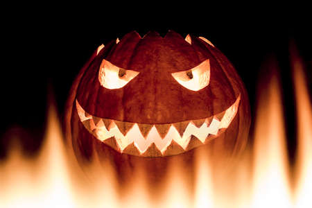 Scary carved halloween pumpkin in hot burning hell fire flames. The big helloween pumpkin has a mad face with glowing eyes and also a glow in its mouth and teeth. Perspective from bottom up.の写真素材