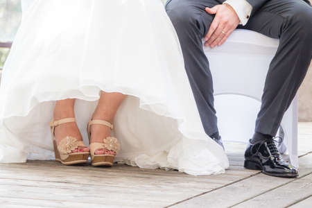 Wedding bride and groom sitting next each other at marriage. The man and the woman wearing luxury bridal dress, suit and shoes. The girl shows her feet with nail varnish. Male shoe is polished.の写真素材