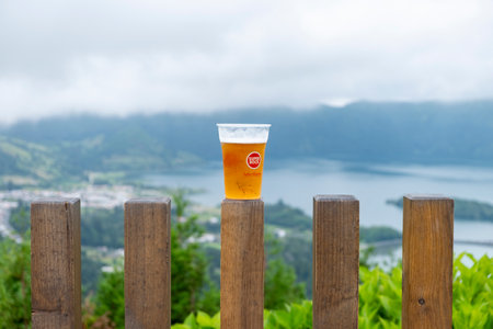 Sao Miguel, Azores - 27.07.2019: Glass of beer of "Super Book - Sabor Autentico" on wooden fence with view of the Seven Cities Lake "Lagoa das Sete Cidades". Copy Space, Close Up.のeditorial素材