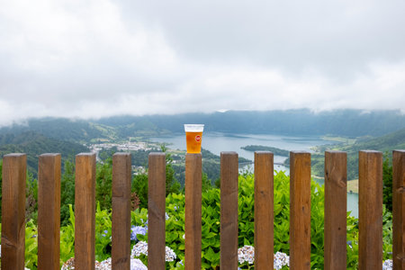 Sao Miguel, Azores - 27.07.2019: Glass of beer of "Super Book - Sabor Autentico" on wooden fence with view of the Seven Cities Lake "Lagoa das Sete Cidades". Copy Space.のeditorial素材