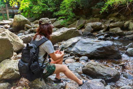 Girl with backpack using smart phone during trekking trip, sitting on the rock near water stream.の写真素材