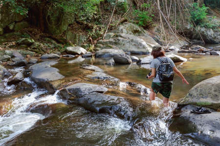 Girl with backpack and camera on trekking trip, walking across the water stream of cascade, during...