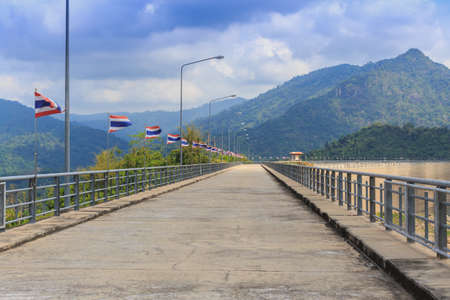 Perspective view of a long road dam, with Thai national flags along the road, mountain and clear blue sky background.の写真素材
