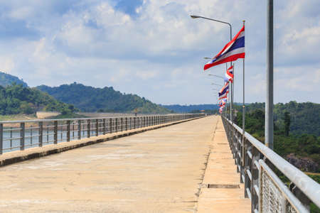 Perspective view of a long road dam, with Thai national flags along the road, mountain and clear blue sky background.の写真素材