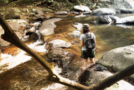 Girl with backpack and camera on trekking trip, walking across the water stream of cascade, during sunbeam shining.の写真素材