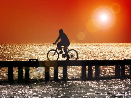 Silhouette of man bicycling across bridge at sunset with lens flareの写真素材
