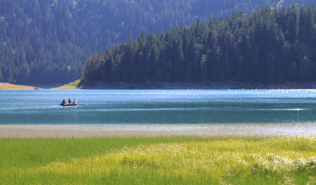 Black Lake in Montenegro. Blured combination of green grass, blue water with small boat and three people inside and darker blue forest covers mountainsの写真素材