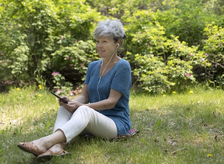 Smiling senior gray-haired woman sitting on green grass and listening to music with tablet computer. Eldery woman uses tablet.の写真素材