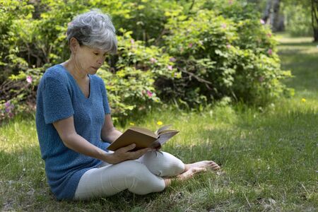 Senior woman reading a book in the garden. Gray-haired woman sitting on the green grass.の写真素材