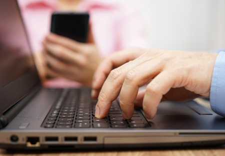 businessman is typing on laptop keyboard with woman in blurred background using mobile smart phoneの写真素材