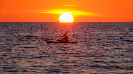 silhouette of man paddling with his canoe in the sea in front of sinking sun just before night timeの写真素材