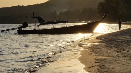 Thai boat Ruea Hang Yao lieing on the beach of Koh Mook with sun reflecting on the wet sand. In the background walks a lonely person along the beach.の写真素材
