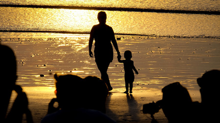 Silhouette of mother and child walking along the beach with sunset mirroring in the sand and people in the front watching the sunsetの写真素材