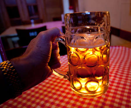 Close up of a liter glass of lager with a foamy head in a man's hand against a red and white tablecloth and backlit by a candleの写真素材