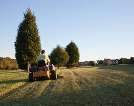 Middle aged man on zero turn mower cutting grass on a sunny day with the sun low in the skyの写真素材