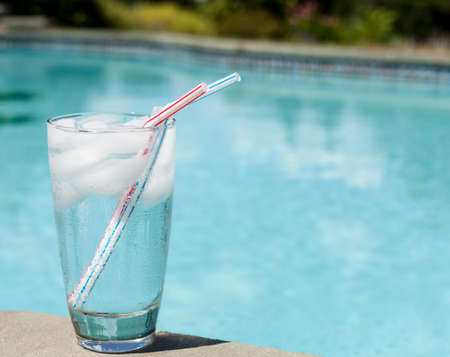 Plain glass of water with ice cubes on side of blue swimming poolの写真素材