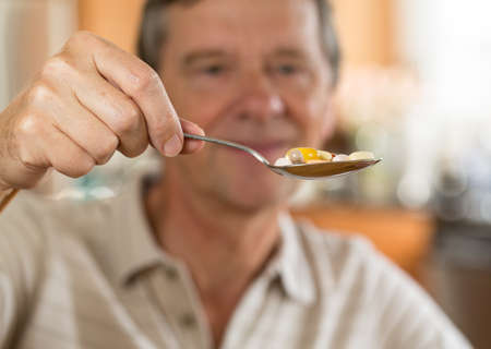 Senior caucasian male eating a spoonful of vitamin tablets for breakfast in kitchenの写真素材