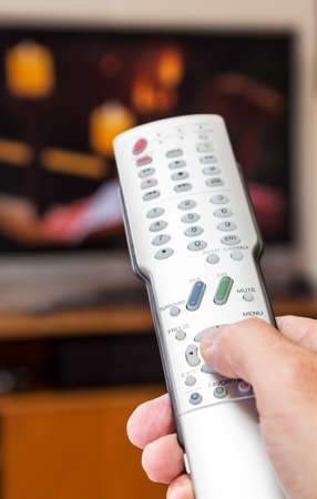 Silver modern TV remote control being pressed by thumb with out of focus screen background