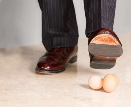 Male brown leather shoe in suit pants about to tread on three brown eggsの写真素材