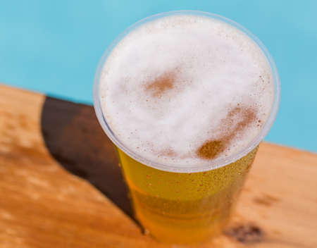 Golden beer, ale or lager a plastic disposable cup for party on wooden table by swimming poolの写真素材