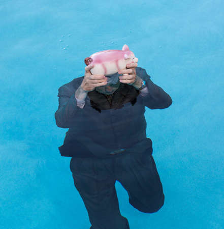 Senior caucasian man holding piggy bank above water as he slowly drowns in debt wearing business suitの写真素材