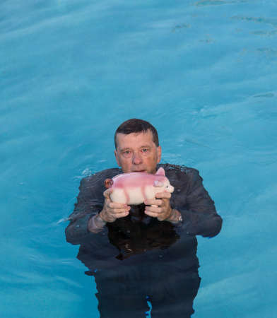 Senior caucasian man holding piggy bank above water as he slowly drowns in debt wearing business suitの写真素材