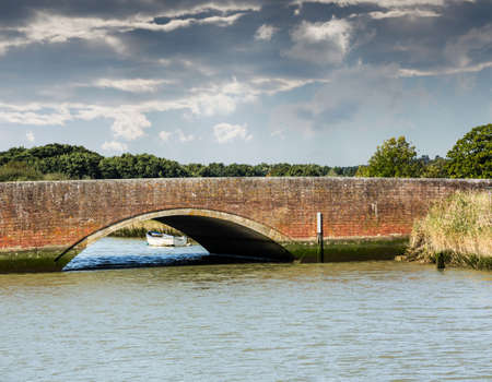 Road arch over Norfolk Broads river with small white rowing boat moored by the bankの写真素材