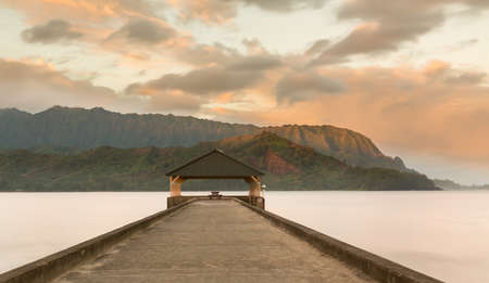 Rising sun illuminates the peaks of Na Pali mountains over the calm bay and Hanalei Pier の写真素材