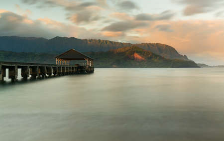 Rising sun illuminates the peaks of Na Pali mountains over the calm bay and Hanalei Pier の写真素材